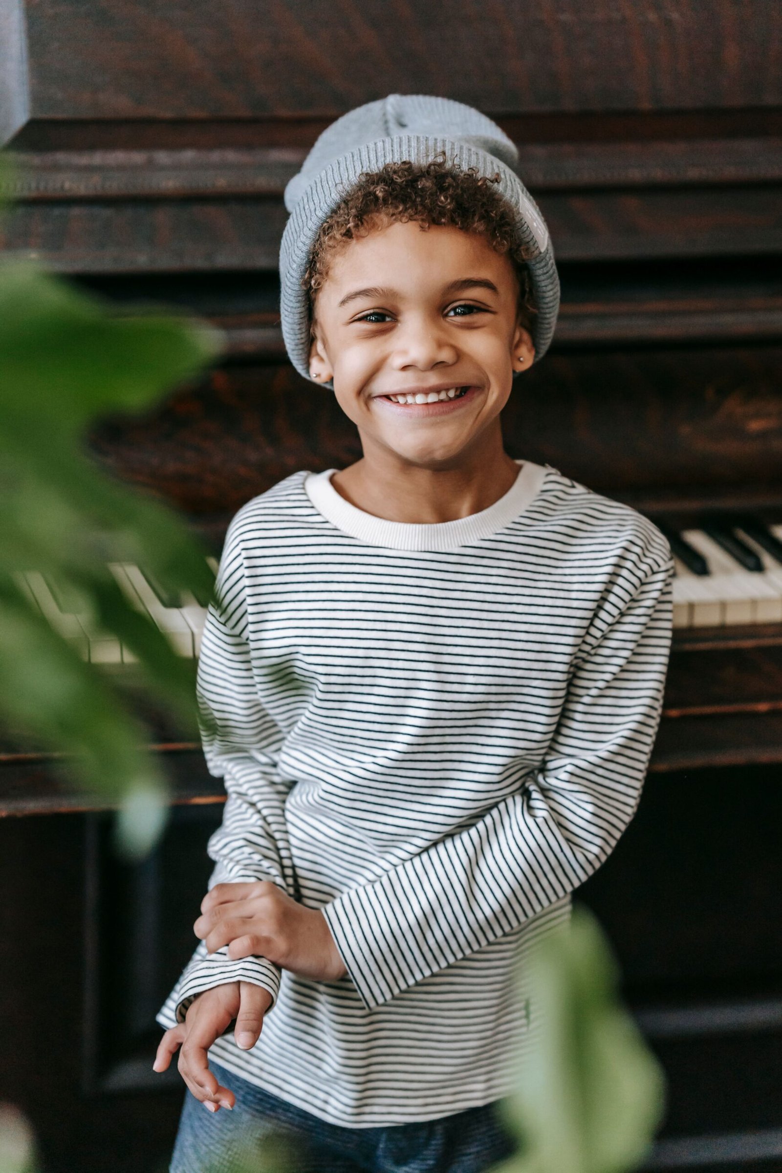 Smiling African American child in hat and casual clothes standing near piano and plant with green leaves at light home and looking at camera